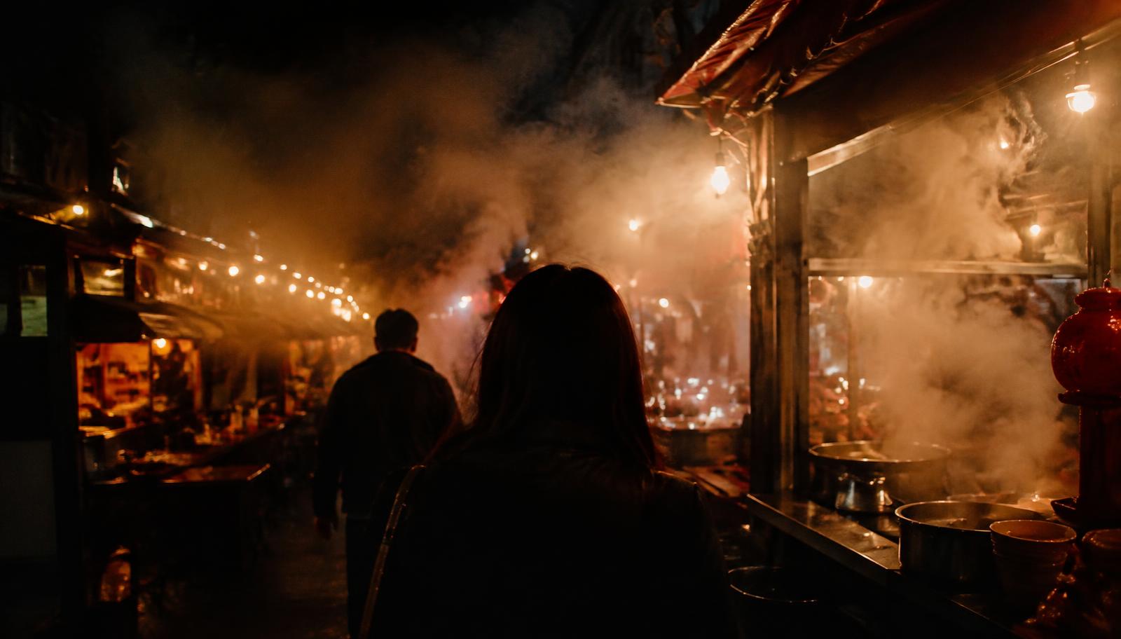 Surreal photorealistic night photography of a Taiwanese night market, an anonymous dreamer following a companion toward a hot pot restaurant, dense steam swallowing strings of warm light bulbs, lively food stalls fading into darkness, intimate melancholy, cinematic documentary framing, realistic textures, no readable text.