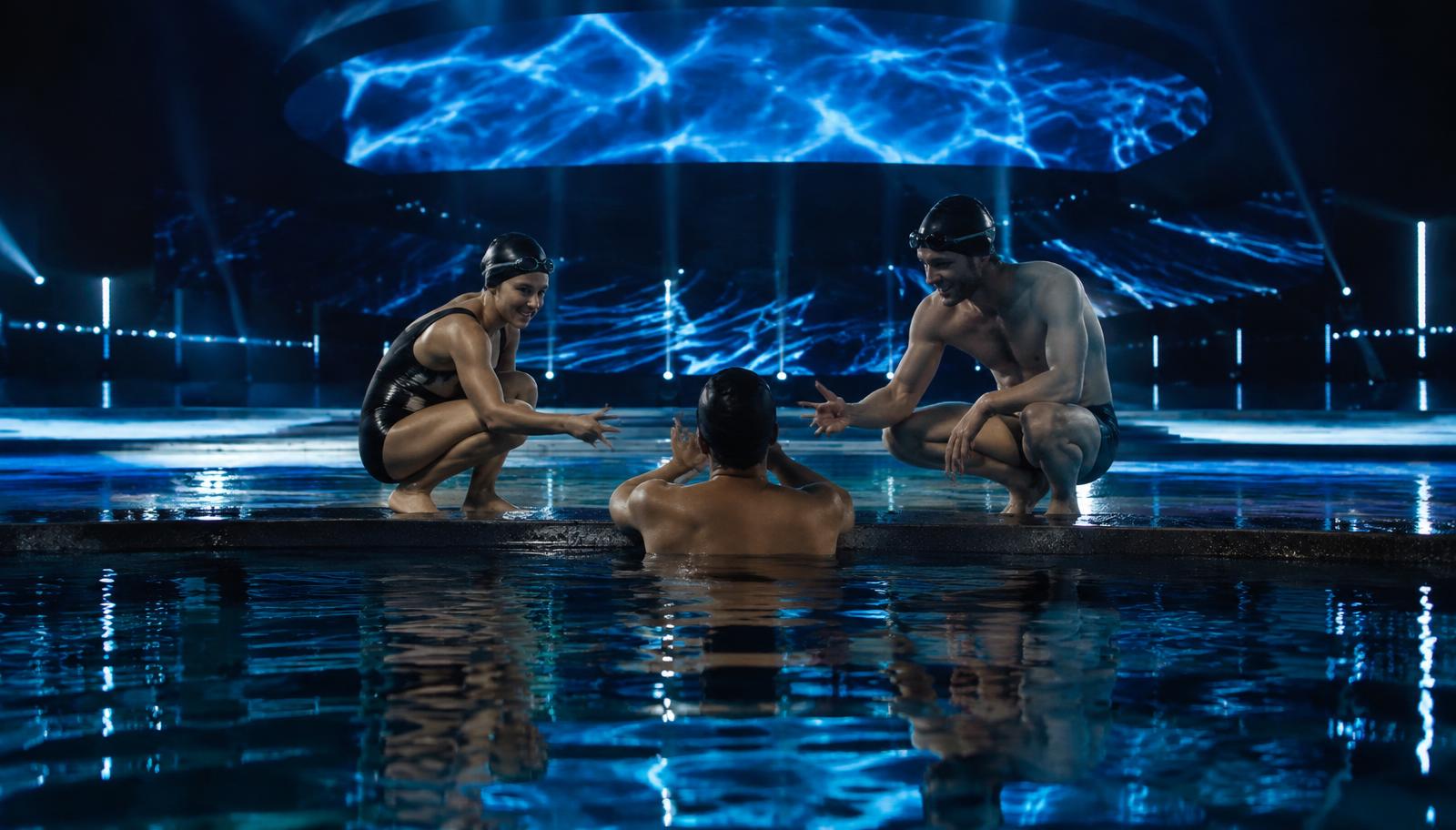 Photorealistic surreal photography of a brightly lit television stage surrounding a cold indoor swimming pool, an anonymous adult swimmer beside the pool edge playing rock paper scissors with two professional adult swimmers, rippling reflections dissolving into darkness, realistic water and skin textures, cinematic wide shot, no text, no lettering, no logos.